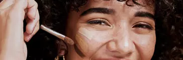 Face, makeup and brush with a woman black model applying foundation to her skin in studio for cosmetics. Portrait, wellness and luxury with an attractive young female using a cosmetic product.